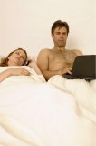Portrait of a man using a laptop in bed while his wife sleeps.