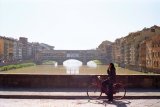 A young woman has stopped her bicycle on the Ponte S. Trinita with the Ponte Vecchio in the background as she talks on her mobile phone.
